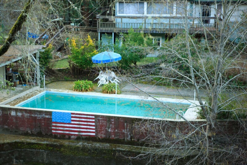 A backyard swimming pool with greenish water, surrounded by bare trees, white poolside chairs, and a blue umbrella. An American flag is displayed on the pool’s retaining wall. A house is visible in the background.