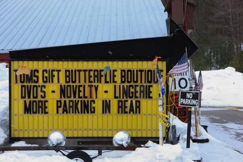 A yellow sign reads “Toms Gift Butterflie Boutique DVD’s - Novelty - Lingerie More Parking in Rear.” Nearby, a “No Parking” sign and American flags stand in snow next to a building with a metal roof.