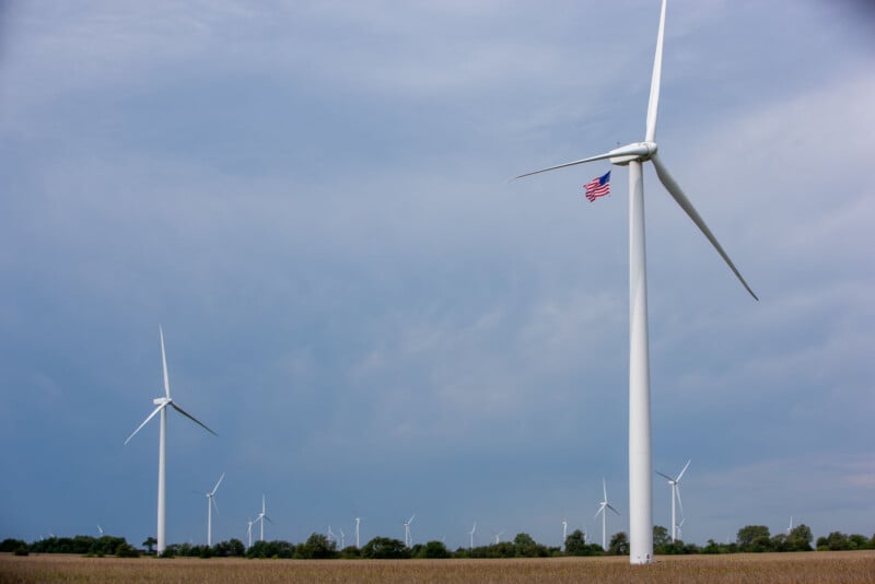 Wind turbines stand in a field under a blue sky, with an American flag attached to the blade of the nearest turbine. Trees and more turbines are visible in the background.