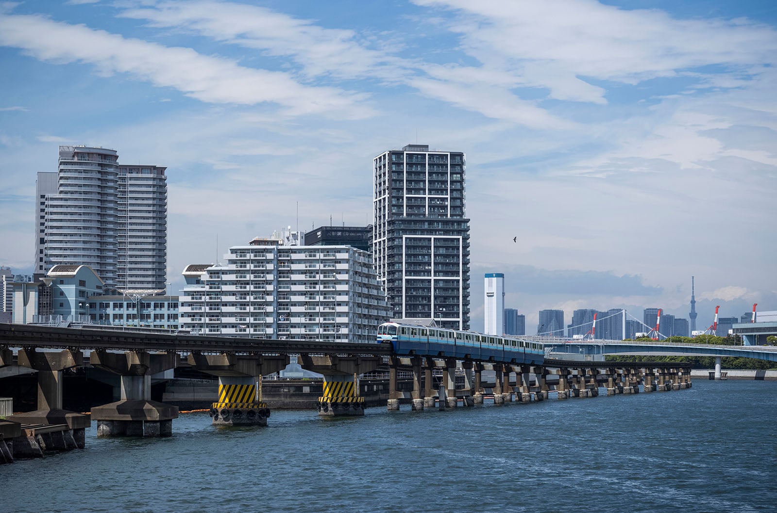A train travels across a bridge over water, with modern high-rise buildings in the background under a partly cloudy sky.