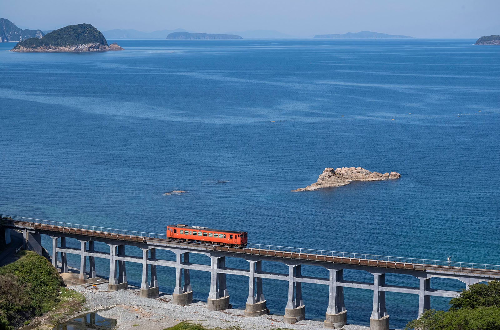 An orange train crosses a long bridge over blue water with small islands and distant hills in the background under a clear sky.