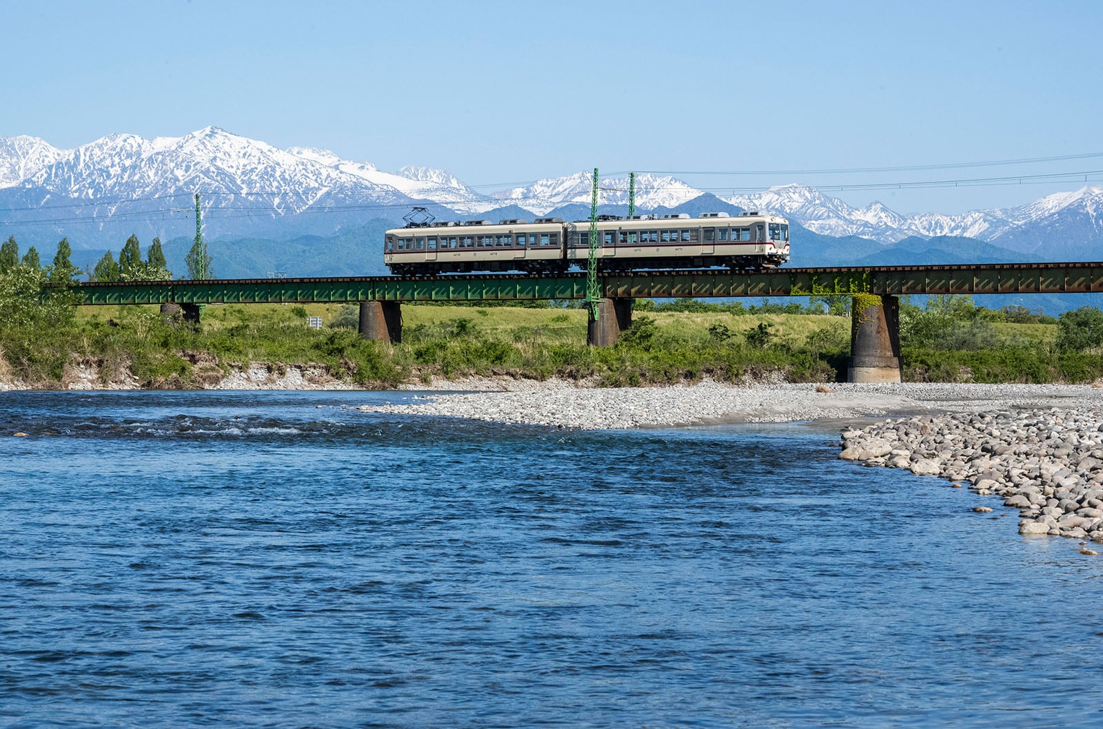 A train crosses a green bridge over a river with rocky banks, with snow-capped mountains and blue sky in the background.
