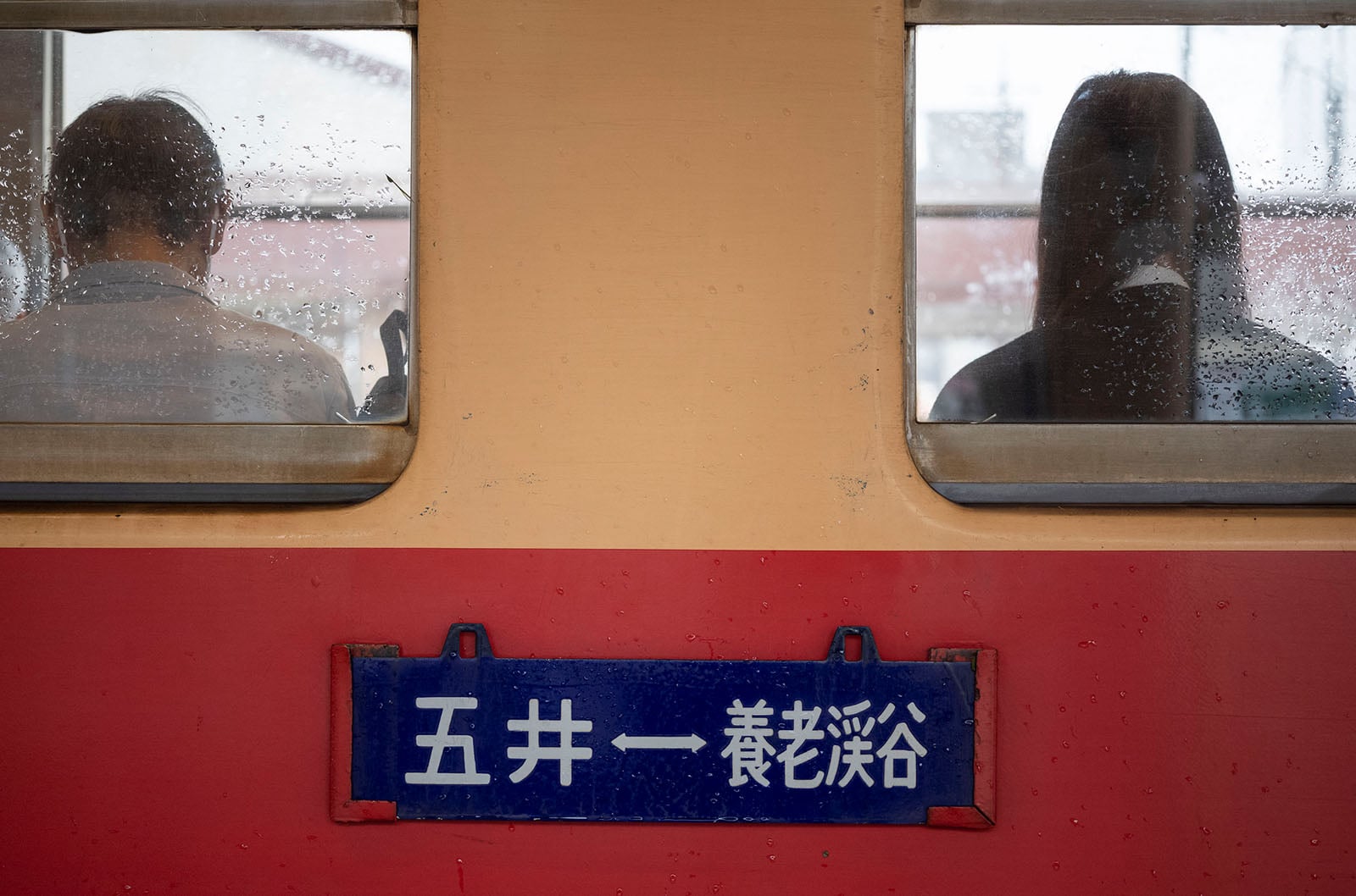 Two people sit back to back, separated by window frames on a rainy train. Raindrops dot the glass, and a blue sign with white Japanese characters is below the windows on the red and beige train exterior.