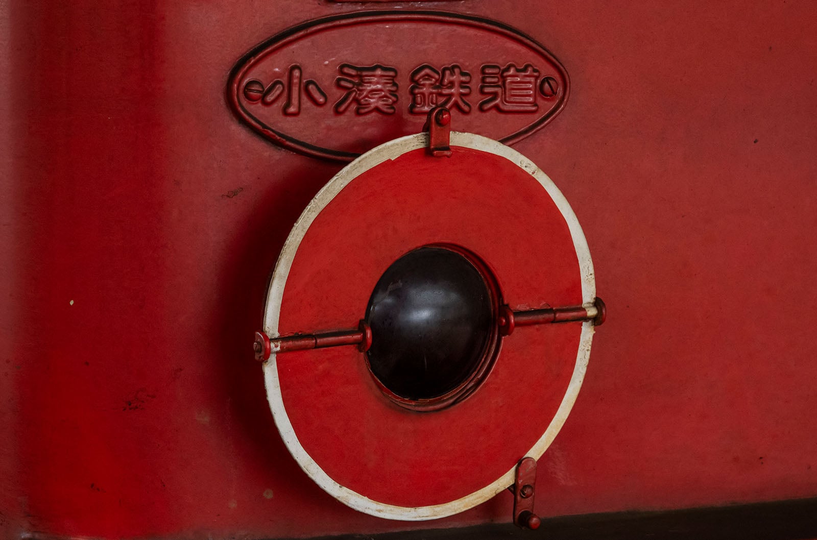 A circular red and white lever with a black center is attached to a red wall. Above it, there is an oval plaque with Japanese characters embossed on it.