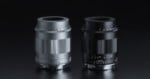 Two camera lenses, one silver and one black, are standing upright side by side on a reflective dark surface, both showing aperture and focus markings.