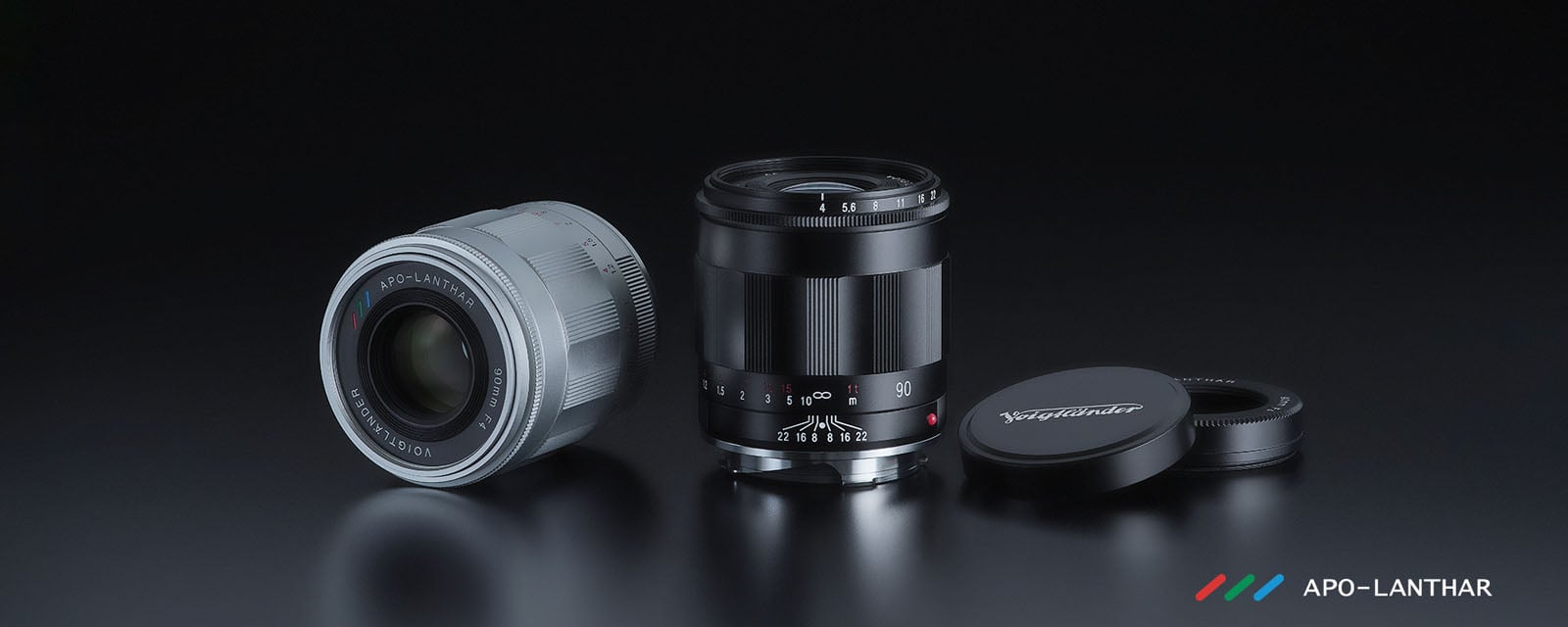Three camera lenses, two standing upright (one silver, one black) and one lying on its side with its front cap off, displayed on a reflective black surface. "APO-LANTHAR" logo is visible in the lower right corner.