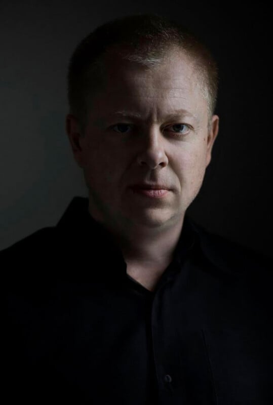 A man with short light hair and fair skin, wearing a black shirt, faces the camera with a serious expression. The lighting is dramatic, illuminating one side of his face while the background remains dark.