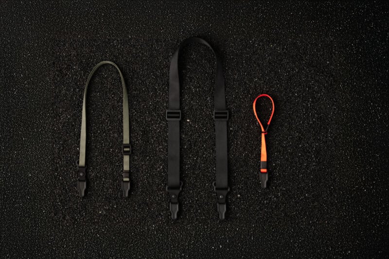 Three camera straps are laid out on a textured black surface: a short black strap on the left, a longer adjustable black strap in the middle, and a smaller orange and black wrist strap on the right.