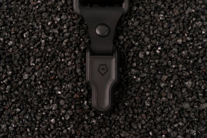 A close-up of a black plastic clip with a geometric logo, resting on a surface covered with small, black, shiny gravel.