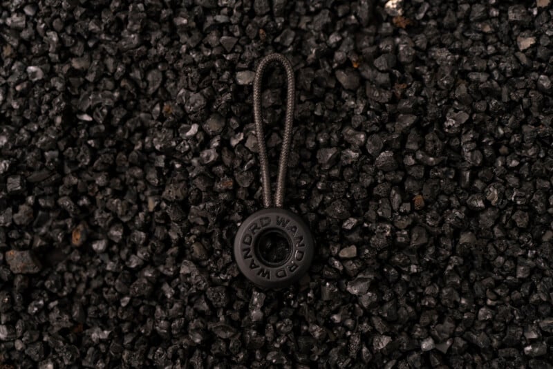 A black keychain with "WANDRD" written on it lies on a surface covered with small, rough, black stones.