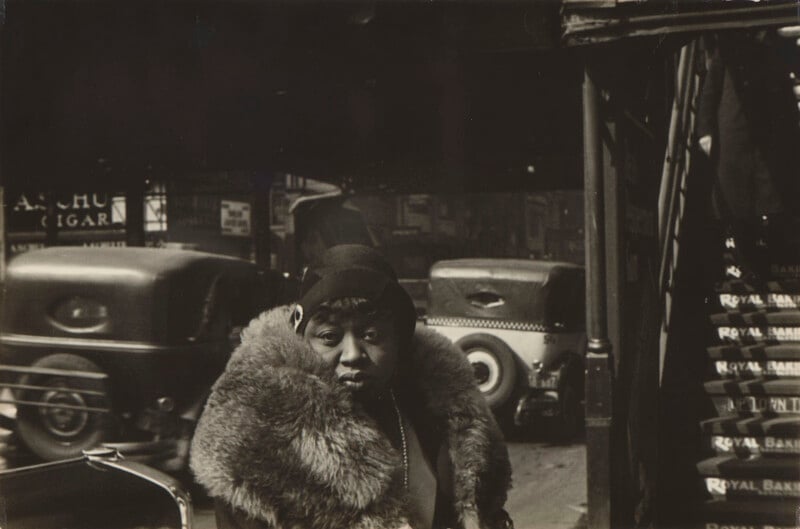 A woman wearing a dark cloche hat and a large fur collar stands on a city street with vintage cars and a staircase in the background.