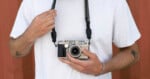 A person in a white t-shirt holds a vintage camera by its strap. Their left arm has a tattoo, and they wear a silver bracelet. The background is a red wall.