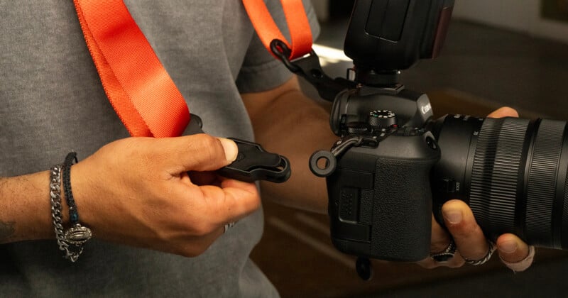 A person attaches an orange camera strap to a black DSLR camera, wearing bracelets and rings on their left hand.