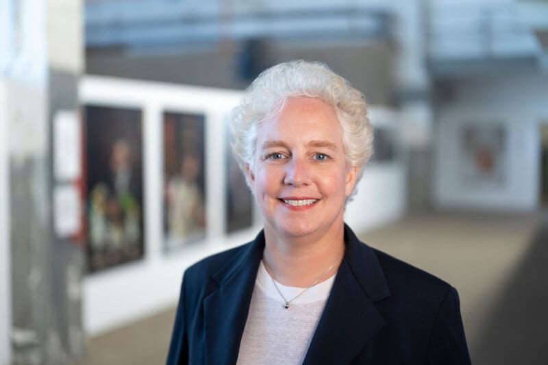 A smiling woman with short white hair wearing a dark blazer and light top stands in a gallery space with blurred artwork in the background.