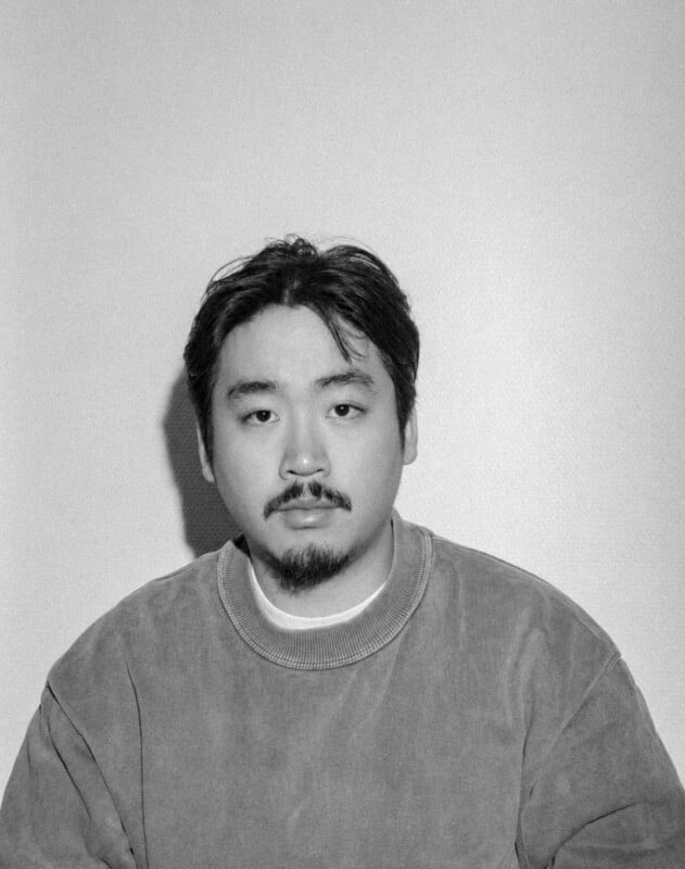 Black and white photo of a man with short dark hair and a beard, wearing a loose sweatshirt, sitting against a plain light-colored wall and looking directly at the camera with a neutral expression.