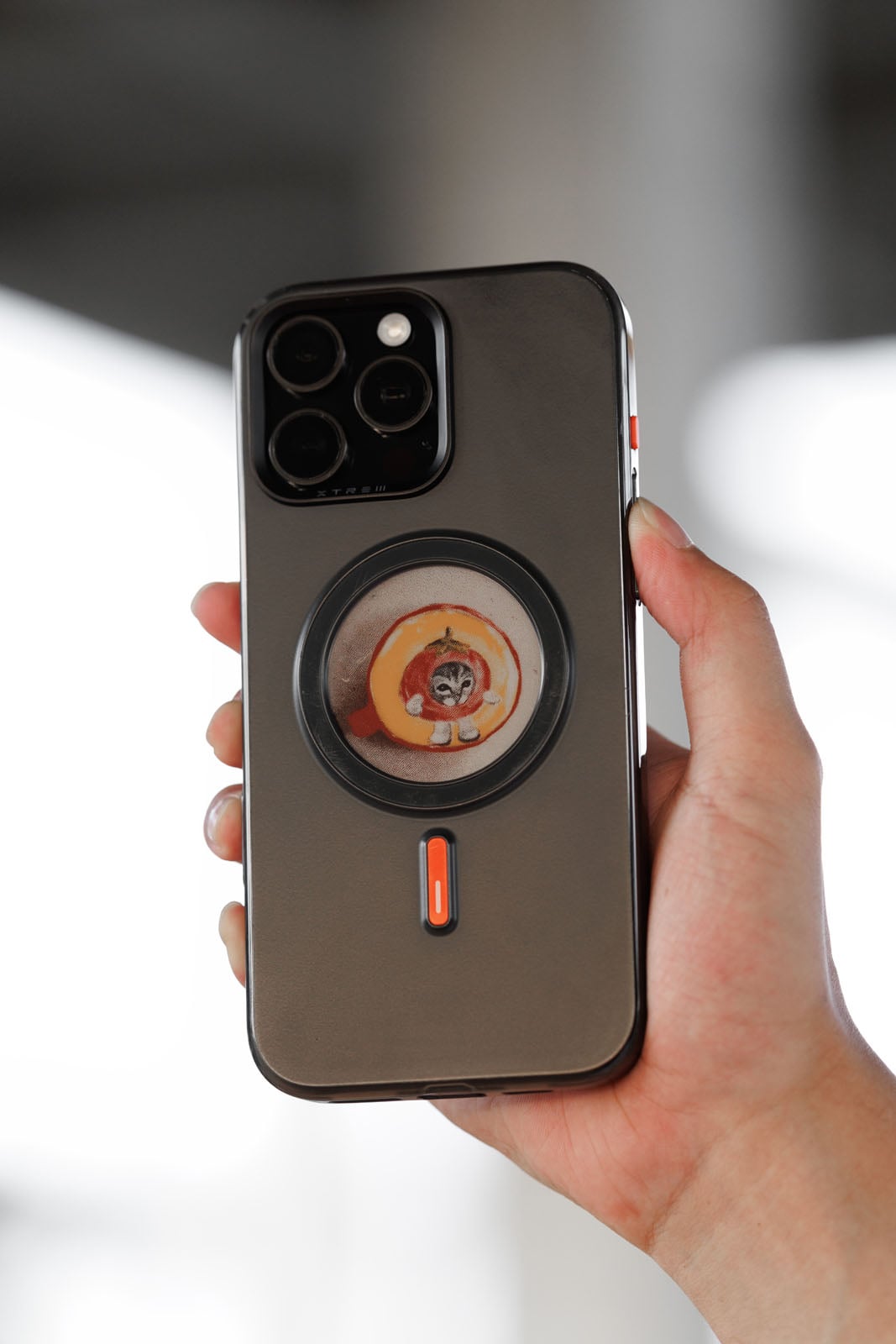 A hand holds a smartphone with a round sticker featuring a cartoon cat inside a peach on the back. The phone case is gray with black edges and an orange button.