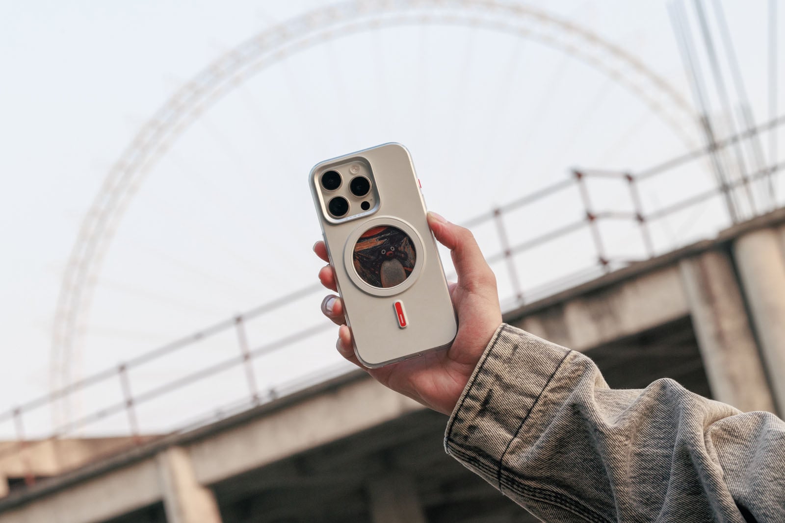 A person in a tan jacket holds up a smartphone with a white case outdoors. A ferris wheel and a concrete structure are visible in the blurred background.
