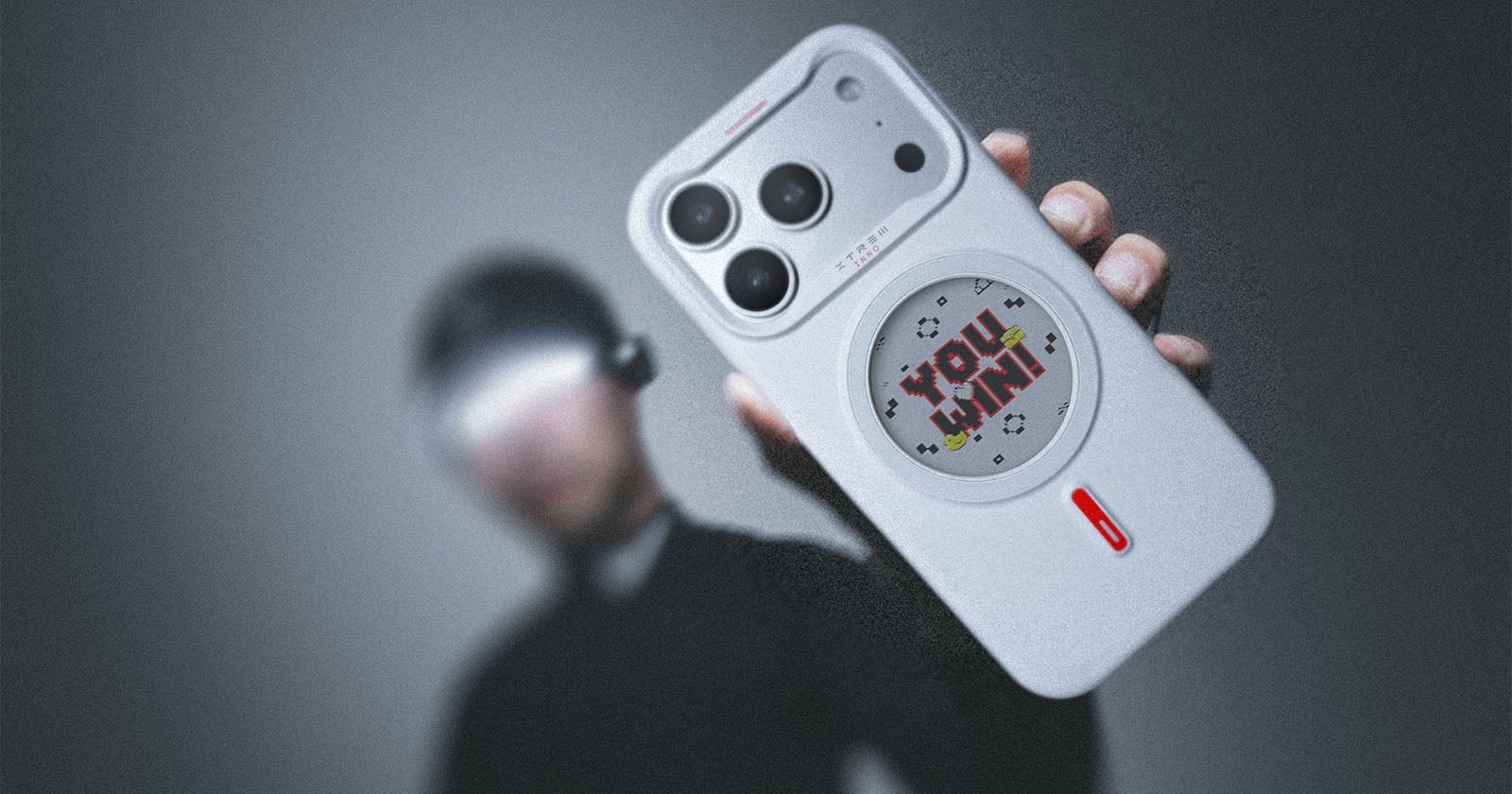 A person holds a white smartphone with large cameras and a sticker that says "YOU WIN!" in pixel art. The background is blurred, and the person's face is obscured.