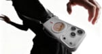 A person wearing black carries a white crossbody bag shaped like a smartphone with camera lenses and a small round screen displaying a cartoon face.