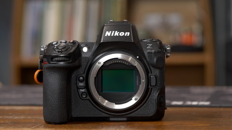 A Nikon Z series mirrorless camera body without a lens, placed on a wooden surface with blurred bookshelves in the background.