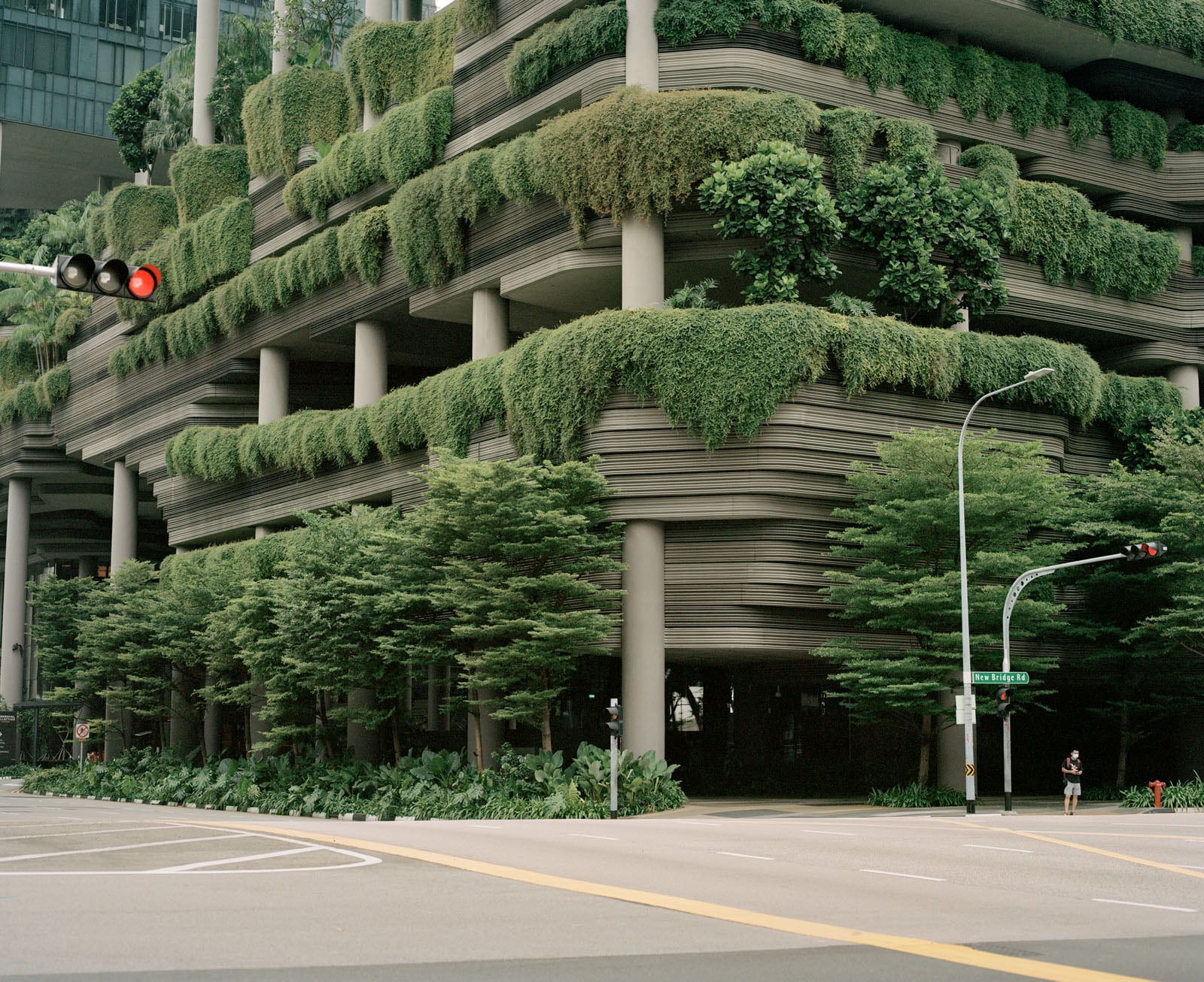 A multi-story building with layered, curved concrete balconies overflowing with lush green vegetation stands at a street corner.