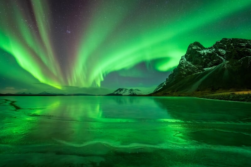 Green northern lights dance across a starry night sky, reflecting over a calm lake with snowy mountains and a rugged, rocky shoreline in the background.