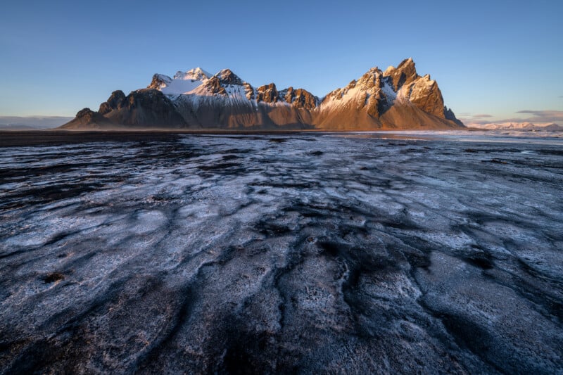 Snow-dusted black sand beach leads to dramatic, jagged mountains under a clear blue sky, with sunlight highlighting the mountain peaks.