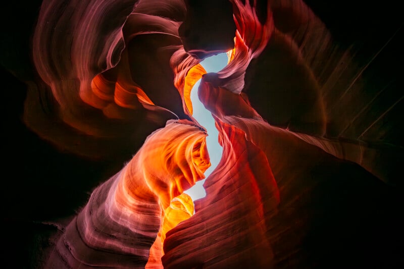 View from inside a colorful slot canyon, showing swirling, wavy sandstone walls in shades of orange, red, and brown, with a sliver of bright blue sky visible through a narrow opening above.
