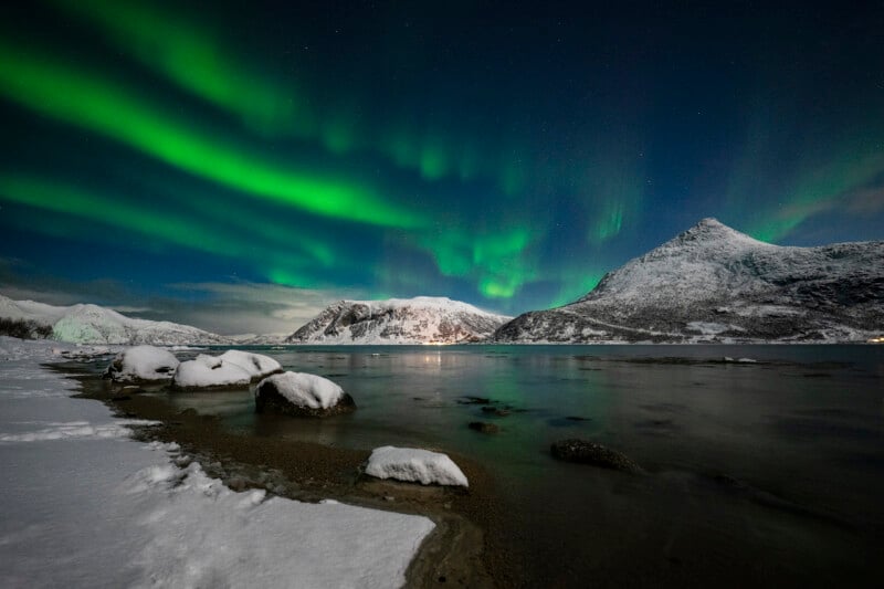 Snowy mountains and shoreline under a night sky with bright green northern lights reflecting on the calm water.