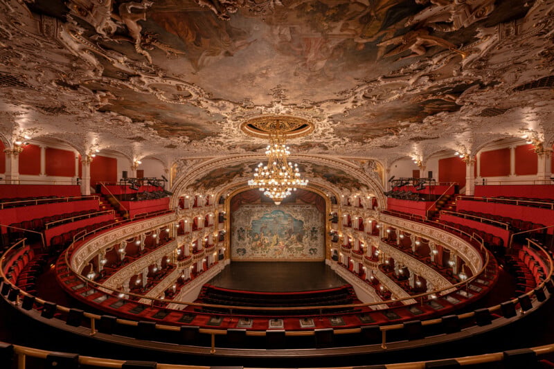 An ornate theater with red seats, golden balconies, a grand chandelier, and a large painted ceiling. The stage features a decorative backdrop, and the architecture is richly detailed and elegant.
