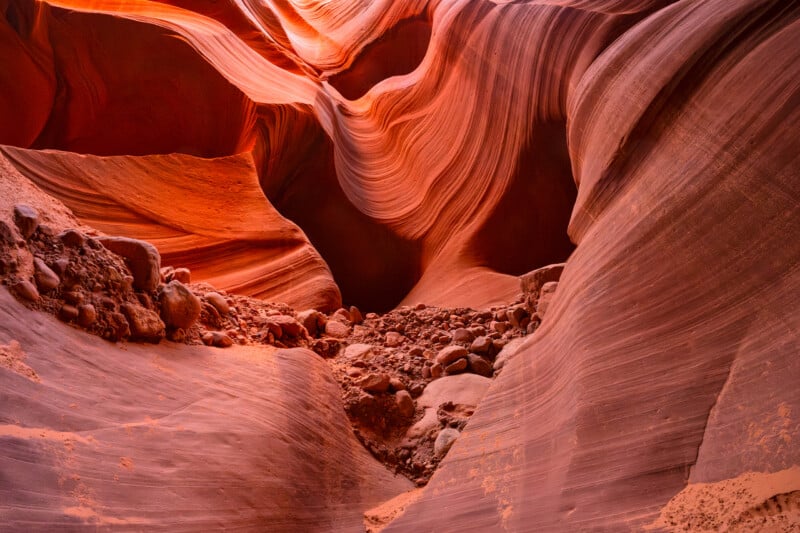 Winding curves and textured walls of a red sandstone slot canyon, with rocks and scattered debris on the canyon floor, lit by warm sunlight filtering through narrow openings above.