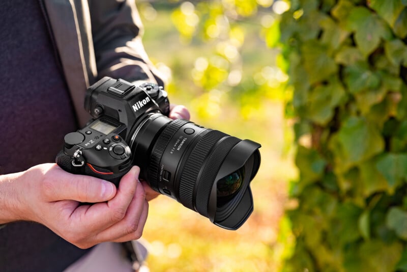 A person holds a Nikon camera with a large lens outdoors near green leaves, preparing to take a photo in bright natural light.