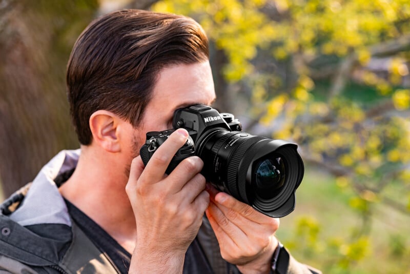 A person holding a Nikon camera up to their face, preparing to take a photo outdoors with greenery and yellow leaves in the blurred background.
