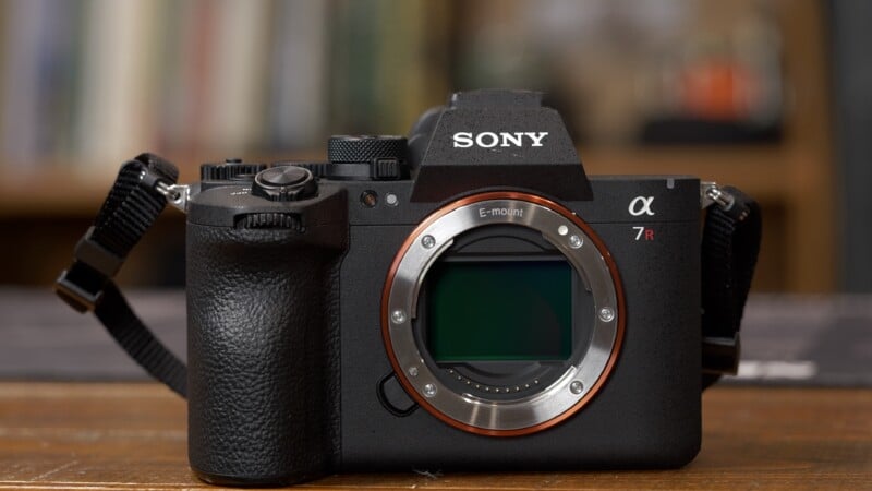 A Sony α7R mirrorless camera without a lens, showing the camera sensor and E-mount. The camera is on a wooden surface with a blurred background.