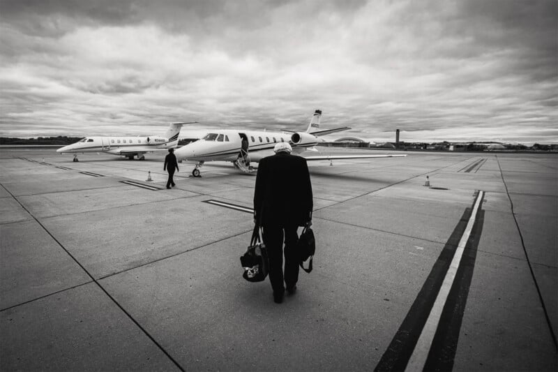 A person carrying bags walks toward a private jet parked on an airport runway, with another jet and figure in the background under a cloudy sky.