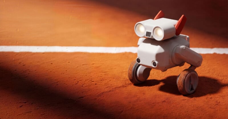 A small, white, robot with two wheels and two red antennae stands on a clay tennis court near a white line, illuminated by sunlight.