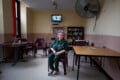 An older woman with purple hair, wearing a green uniform, sits alone in a sparsely furnished room with chairs, tables, bookshelves, and a TV mounted on the wall.