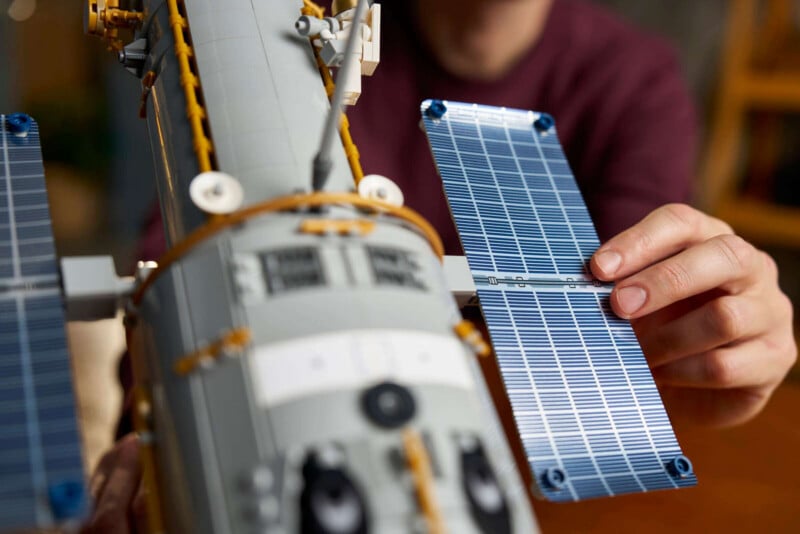 A person attaches a blue solar panel piece to a gray LEGO spacecraft model, focusing on the detailed assembly with their hand visible in the foreground.