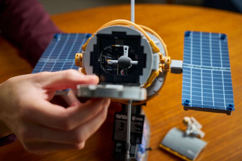 A hand holds part of a detailed LEGO satellite model with blue solar panels, positioned on a wooden surface.
