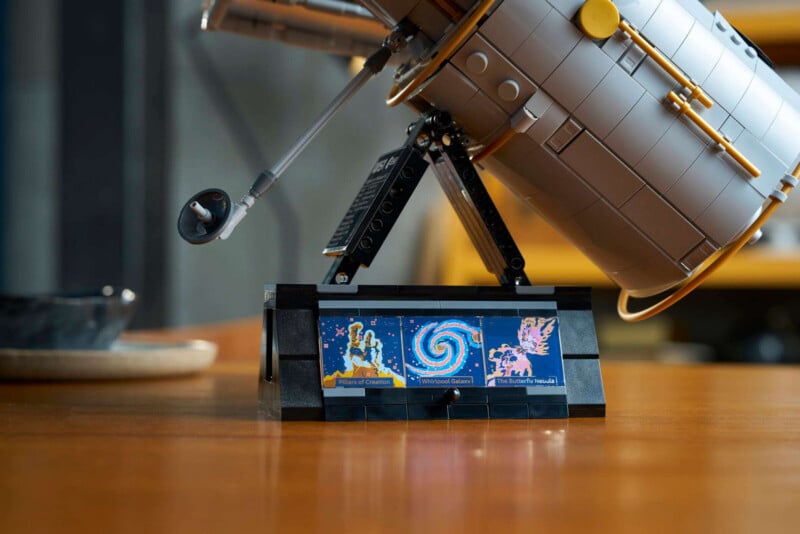 A LEGO model of the Hubble Space Telescope sits on a wooden table with a display stand showing three colorful space-themed tiles.