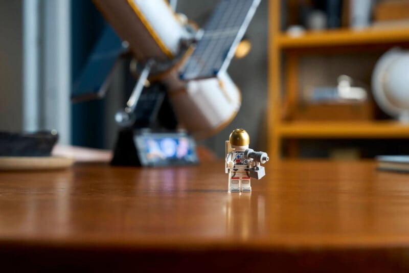 A LEGO astronaut minifigure stands on a wooden table with a blurry model of a space telescope and shelves in the background.
