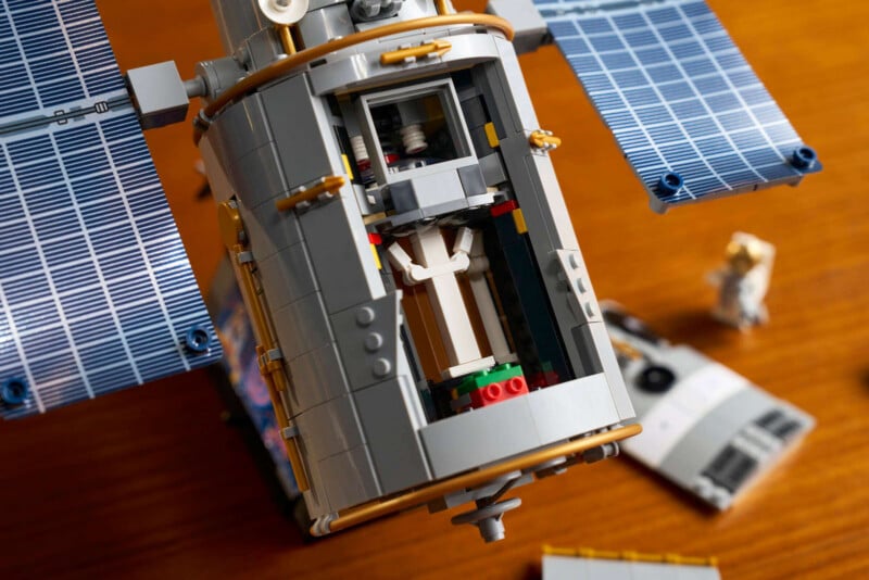 Close-up of a LEGO space satellite model with solar panels, showing an open compartment. Nearby, LEGO astronaut figures and space station parts are on a wooden surface.