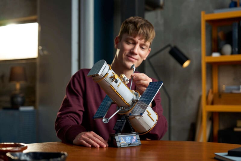 A person smiles while assembling a detailed model of a space telescope on a wooden table in a room with shelves and a lamp in the background.