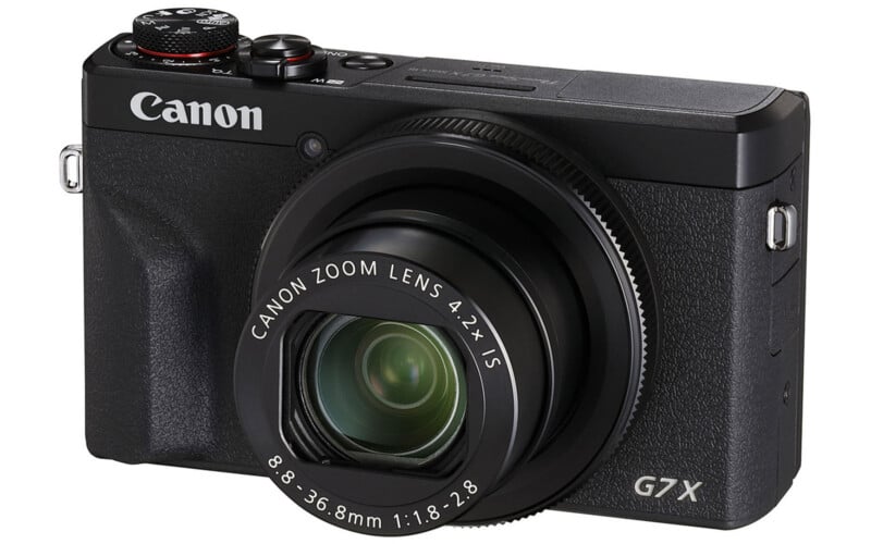 A black Canon G7X digital camera with a zoom lens extended, viewed from the front-left angle against a white background.