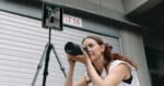 A woman holding a camera prepares to take a photo, next to a tripod-mounted tablet displaying a live preview of her shot, in front of a metal garage door.