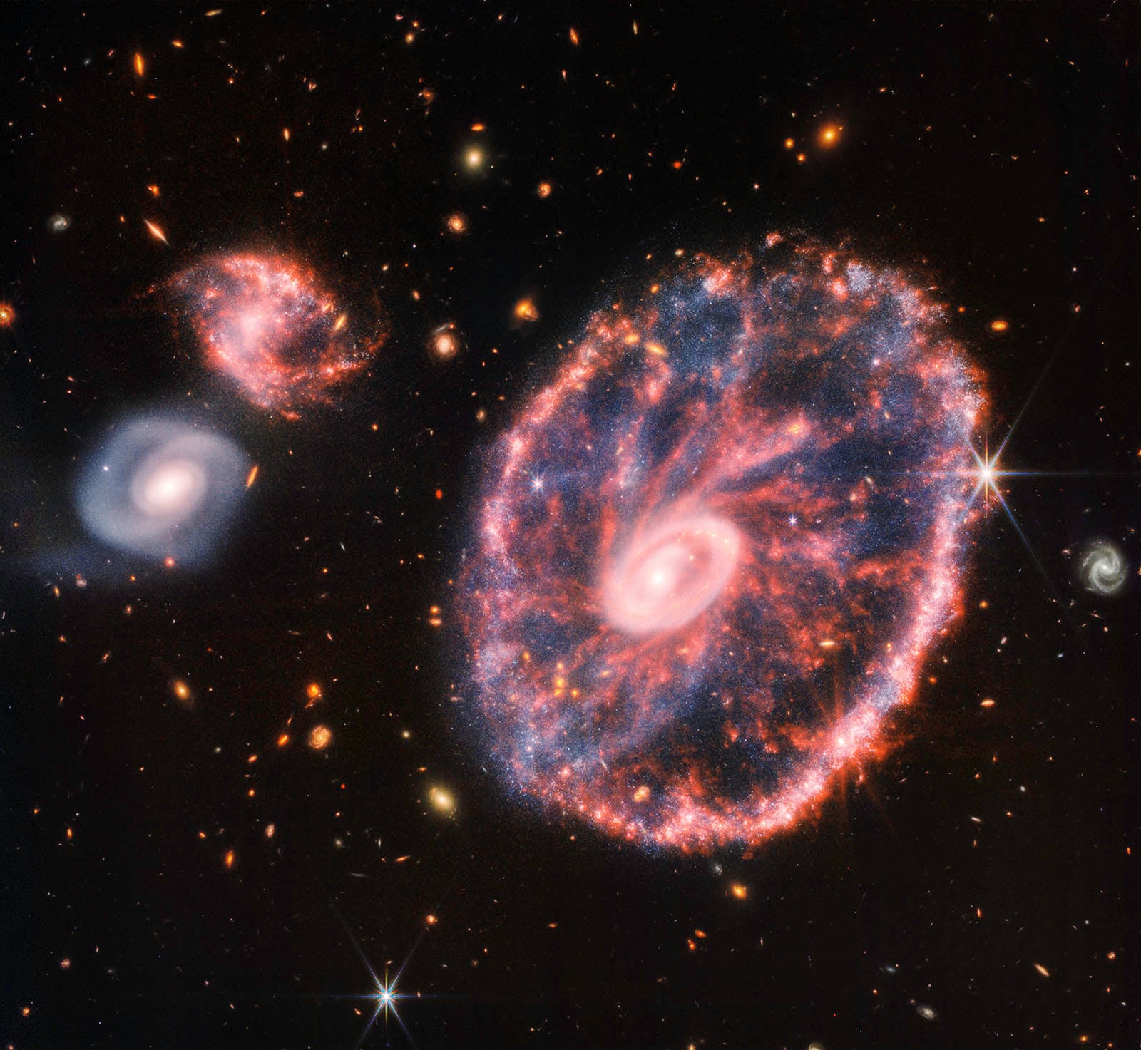 A colorful galaxy with a bright core and ring structure is surrounded by smaller galaxies and stars against a black space background. The main galaxy glows with blue, pink, and white tones.