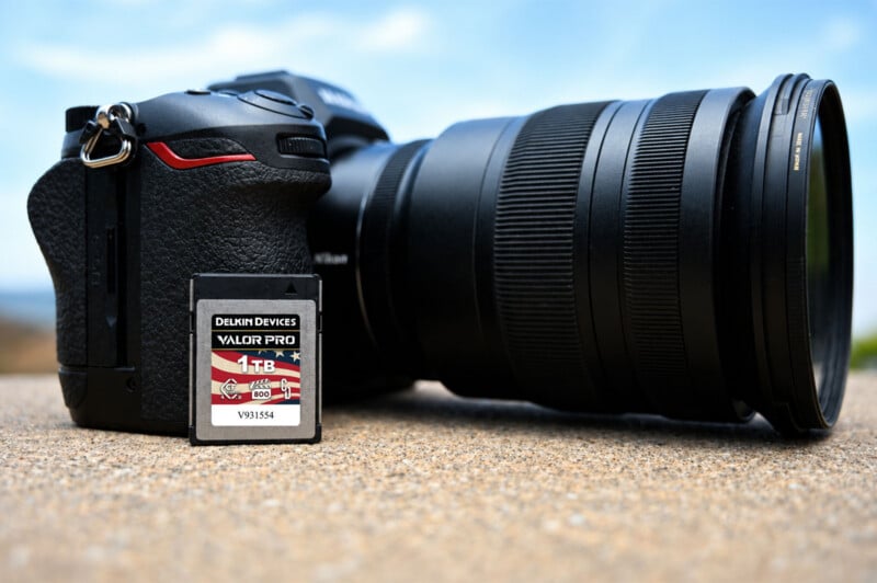 A digital camera with a large lens is positioned on a sandy surface outdoors, with a Delkin Devices VALOR PRO 1TB SD memory card placed upright in front of it.