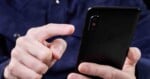 A person in a dark shirt is holding a black smartphone in one hand and using their other hand to tap the screen.