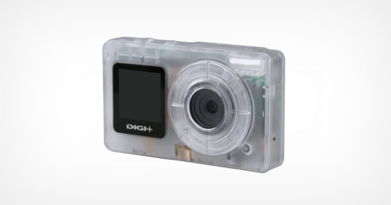 A compact digital camera with a transparent plastic case, revealing internal components. The front features a lens on the right and a small display screen labeled 