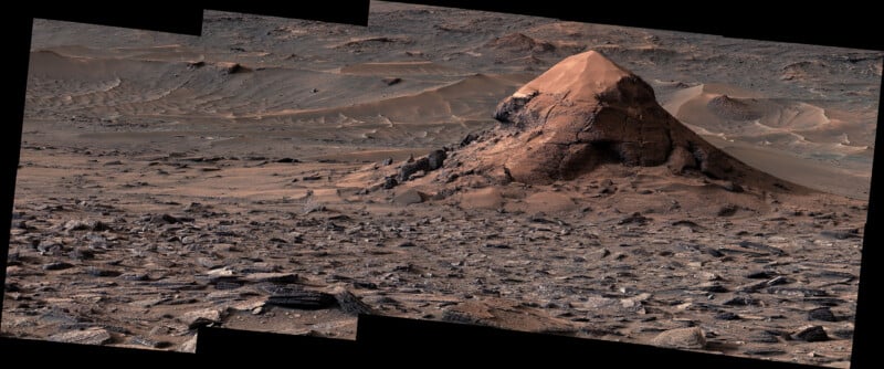 A rocky Martian landscape features a prominent, cone-shaped hill surrounded by sand dunes and scattered, layered rock fragments.